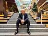 Eivind Roald sits on broad steps inside a modern, plant-filled building with a Christmas tree behind him.