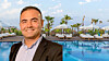 Smiling businessman in a blazer standing by a luxury resort pool with palm trees and loungers.