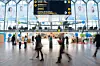 Blurred passengers walk through a bright terminal beneath a large electronic information board.