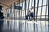 Two travellers walk through a sunlit airport terminal with large glass windows and blue signage.