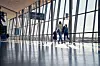 Two travellers walk through a sunlit airport terminal with large glass windows and blue signage.
