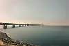 Lång bro över lugnt vatten en disig dag med stenig strand i förgrunden.