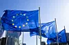 EU-flaggor vajar framför glasbyggnader mot en klar blå himmel.
