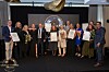 Ryhmä ihmisiä seisoo lavalla gaalatilaisuudessa ja pitää palkintodiplomeja sekä kukkakimppuja.