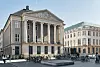 Stor klassisk byggnad med kolonner vid ett öppet torg med cyklar och några personer.