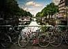 Cyklar står parkerade vid en kanal med båtar och träd längs vattnet i Amsterdam.