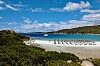 Solstolar i rader på en sandstrand vid turkost vatten, med gröna kullar och blå himmel i bakgrunden.