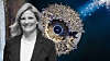 Smiling woman in a blazer stands beside a glowing circular sculpture on a dark blue background.