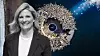 Smiling woman in a blazer stands beside a glowing circular sculpture on a dark blue background.