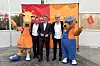 Three men pose with two orange and blue mascots in front of branded event displays.