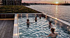 People in a pool beside a waterfront at sunset, with city skyline and warm reflections on the water.
