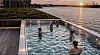 People in a pool beside a waterfront at sunset, with city skyline and warm reflections on the water.