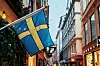 En svensk flagga hänger på en gata i Gamla stan, Stockholm med butiksfönster och byggnader i varmt ljus.