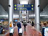 Resenärer med rullväskor går genom en flygplatsterminal med skyltar mot avgångar och incheckning.