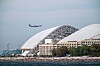 Ett passagerarflygplan flyger över en flygplats med stora välvda tak; vatten syns i förgrunden.