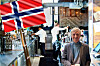 Man standing in a busy bar beneath a Norwegian flag, with staff working behind the counter.