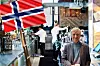 Man standing in a busy bar beneath a Norwegian flag, with staff working behind the counter.