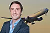 Leende man i mörk kostym framför ett passagerarflygplan i luften.