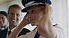 Naval officer adjusting a peaked cap as colleagues smile behind him.