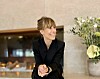Leende kvinna i svart klädsel sitter i en hotellmiljö med en blomsterbukett bakom sig.