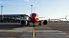 Rött och vitt passagerarflygplan sett framifrån på en flygplats med terminal och blå himmel i bakgrunden.