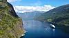 A white cruise ship sails through a deep blue fjord between steep, green mountains.