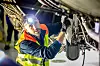 Aircraft technician wearing a high-visibility vest inspects equipment beneath an aircraft.